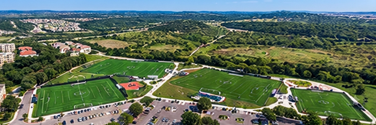Aerial view of a soccer field surrounded by trees, buildings, roads, vehicles, and grass
